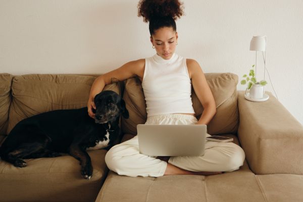 mulher sentada no sofá, um cachorro deitado também e um notboock no seu colo.